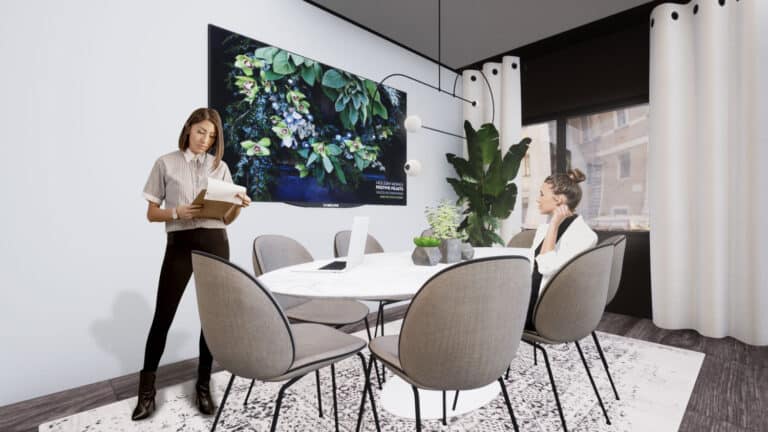 Kokoushuone, jossa soikean pöydän ympärillä on pehmustettuja tuoleja. Seinällä on tv, ja kulmassa on korkea viherkasvi.