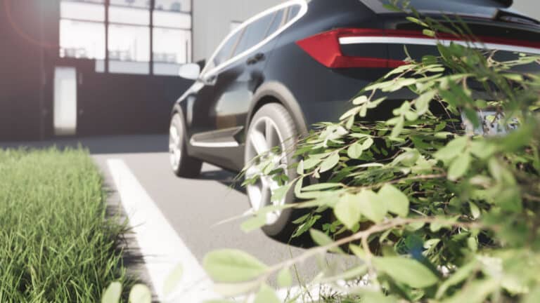 Musta auto pysäköitynä Höyläämön edessä. Parkkipaikan vieressä on vihreää nurmea, ja etualalla on pensaan vehreitä oksia.